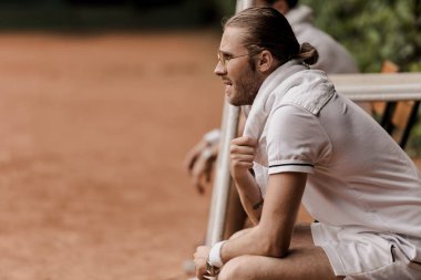 side view of retro styled tennis player sitting on chair at tennis court and looking away clipart