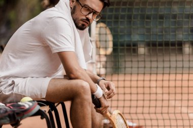 Tenis raketi ile sarayda iskemlede ve kameraya bakarak ciddi tenis oyuncusu