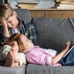 Petite fille couchée avec la tête sur les jambes du frère et en utilisant un ordinateur portable pendant qu'ils se détendre sur le canapé