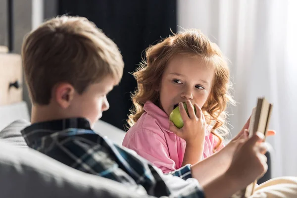 Carino Piccolo Studente Lettura Libro Divano Mentre Sorella Mangiare Mela — Foto Stock