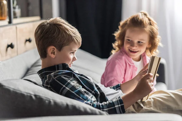 Cute Little Uczniak Czytanie Książki Kanapie Jego Siostra Mówić Niego — Zdjęcie stockowe