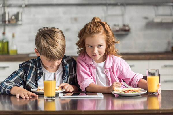 cute little brother and sister using tablet during breakfast at kitchen