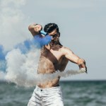 Atractivo hombre sin camisa bailando con palos de humo azul y blanco en frente del océano