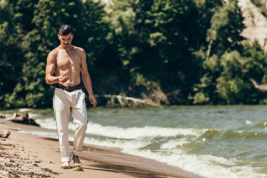 handsome shirtless man looking at his hand and walking by sandy beach clipart