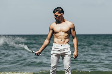 handsome shirtless man holding white smoking stick in front of sea clipart