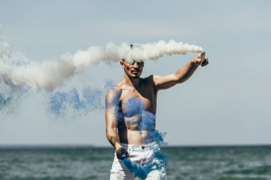 attractive man holding blue and white smoke sticks in front of sea clipart