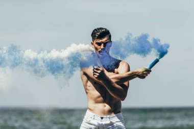 muscular shirtless man with blue and white smoke sticks in front of ocean clipart