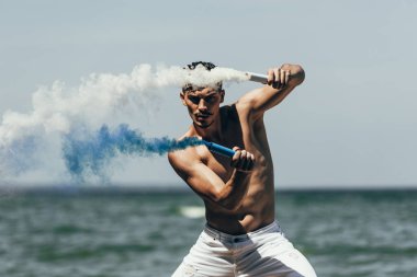 muscular shirtless man dancing with blue and white smoke sticks in front of ocean clipart