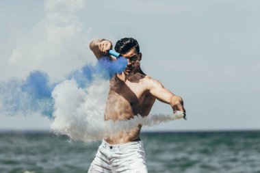 attractive shirtless man dancing with blue and white smoke sticks in front of ocean clipart
