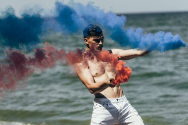 shirtless man dancing with red and blue smoke sticks in front of ocean view clipart