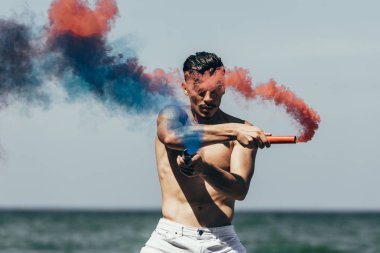 handsome shirtless man with red and blue smoke sticks in front of ocean view clipart