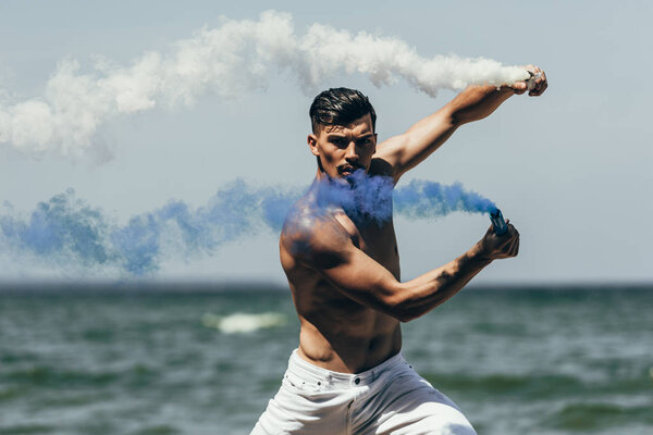 handsome shirtless man dancing with blue and white smoke sticks in front of ocean