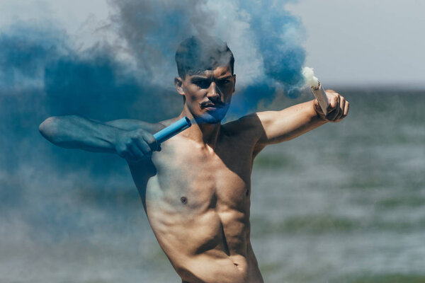handsome shirtless man with blue and white smoke sticks in front of ocean