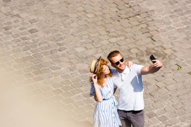 high angle view of smiling redhead man in sunglasses taking selfie with girlfriend on smartphone at city street clipart