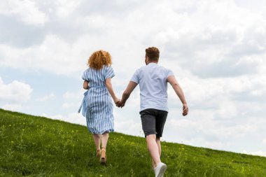 Elele ve çimenli tepeye çalışan kızıl saçlı çift arka görünüm 