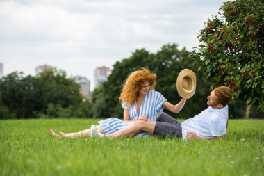 smiling redhead woman trying to putting on own straw hat on boyfriend head on grass in park  clipart