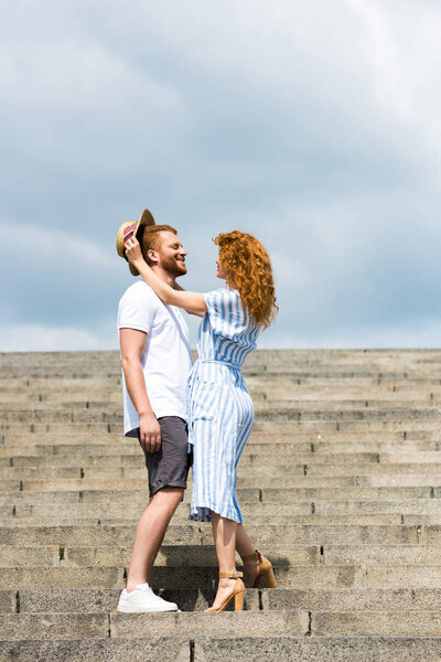 attractive redhead woman putting on own straw hat on boyfriends head on stairs 
