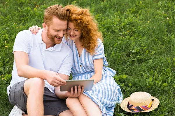 smiling redhead couple with digital tablet on grassy meadow
