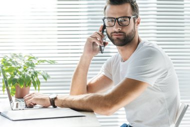 brunet yakışıklı dizüstü bilgisayar kullanırken Smartphone'da konuşuyor