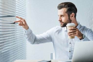 kahve tutarak ve jaluzi pencereden bakarak işadamı