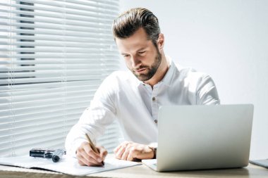 concentrated businessman writing in documents while sitting at workplace with laptop and gun