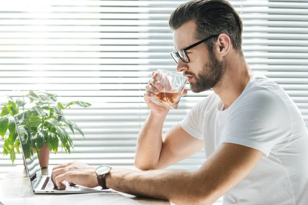 Schöner Bärtiger Mann Trinkt Whiskey Während Laptop Arbeitsplatz Benutzt — Stockfoto