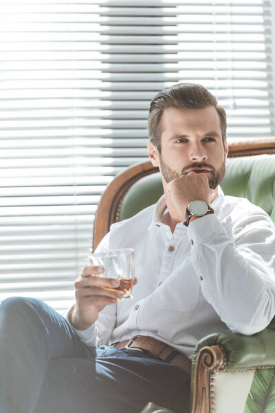 бородатый задумчивый мужчина держит стакан коньяка и сидит в кресле
