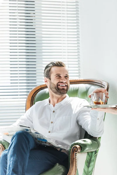 handsome smiling man with newspaper taking whiskey glass from tray