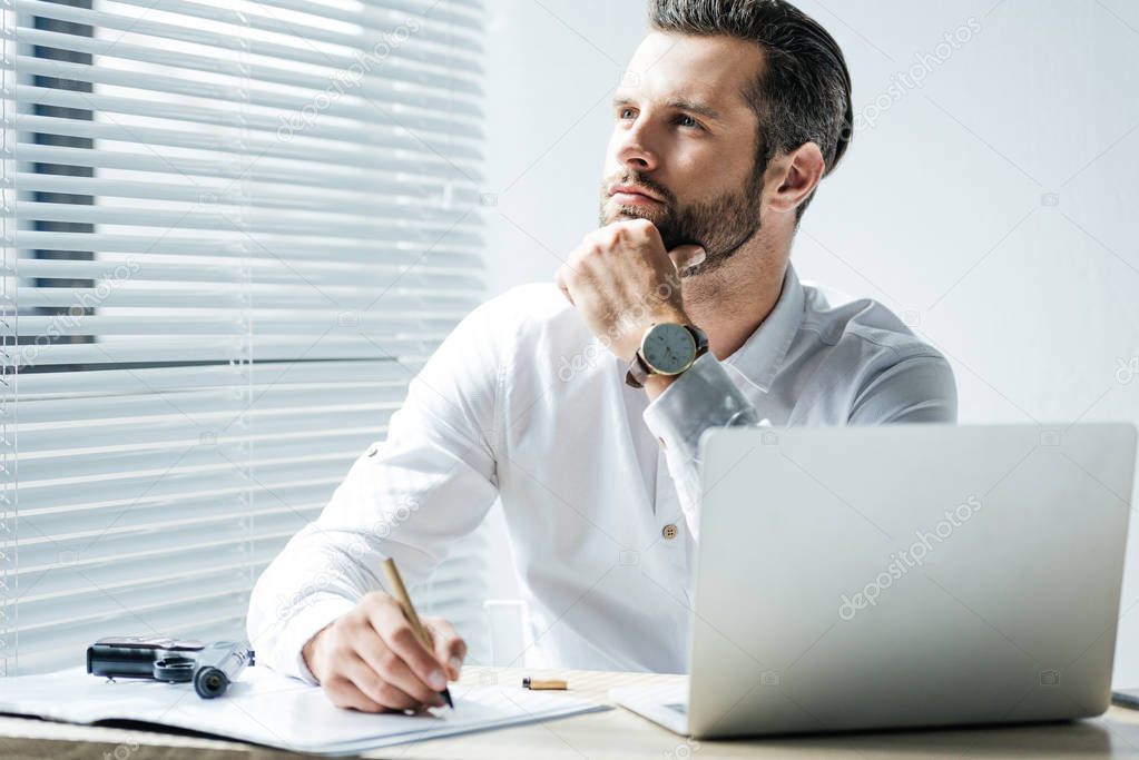 pensive businessman doing paperwork in office with laptop and gun