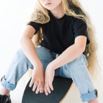 High angle view of child with long curly hair sitting on skateboard and looking at camera isolated on white