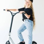 Full längd syn på barn i svart t-shirt och mössa ridning scooter och tittar kameran isolerad på vit