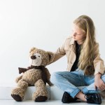 Vista de longitud completa de lindo niño elegante sentado y mirando oso de peluche en blanco