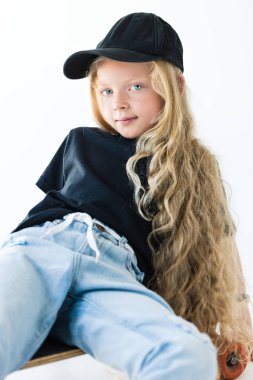 kaykay üzerinde oturan ve üzerinde beyaz izole kamera gülümseyen uzun kıvırcık saçlı sevimli çocuk