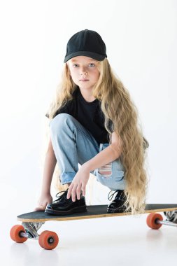 kaykay üzerinde çömelmiş ve beyaz kameraya bakarak uzun kıvırcık saçlı güzel çocuk