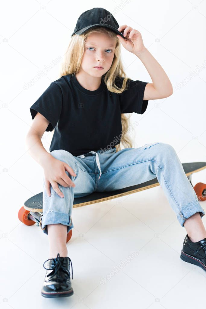 high angle view of kid in black cap and t-shirt sitting on longboard and looking at camera isolated on white