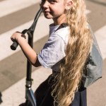 Высокий угол зрения красивой маленькой школьницы с длинными кудрявыми волосами, катающейся на скутере и смотрящей в камеру