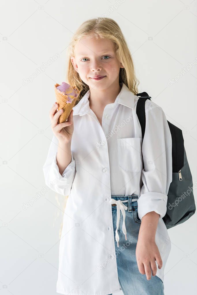 adorable child with backpack eating ice cream and smiling at camera isolated on white
