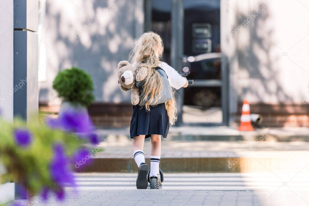rear view of adorable schoolchild with backpack and teddy bear riding scooter on street   