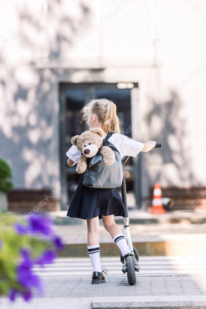 back view of adorable little schoolgirl with backpack and teddy bear riding scooter on street  
