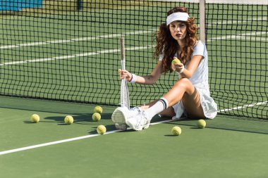 beautiful female tennis player with racket sitting near tennis net on court with tennis balls around clipart