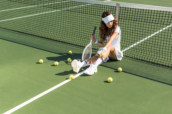 beautiful female tennis player with racket sitting near tennis net on court with tennis balls around