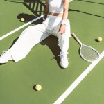 Young woman in fashionable white clothing and cap sitting on tennis court with racket and balls