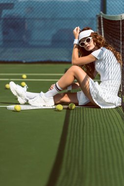 fashionable female tennis player in sunglasses resting near net on tennis court with equipment near by   clipart