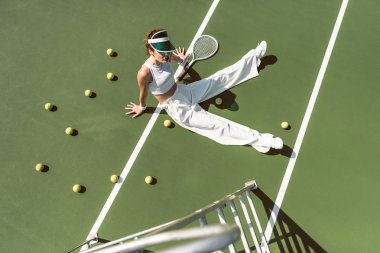 high angle view of beautiful woman in stylish white clothing sitting on tennis court with balls and racket around clipart