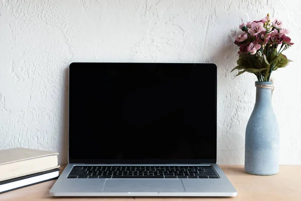 laptop with blank screen, flowers in vase and books on table