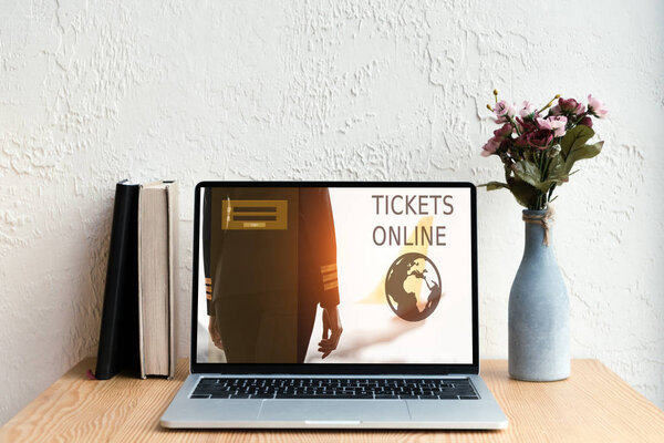 laptop with tickets online website on screen, books and flowers in vase on wooden table