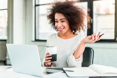 kahve tutarak ve office içinde laptop bakarak mutlu Afro-Amerikan işkadını