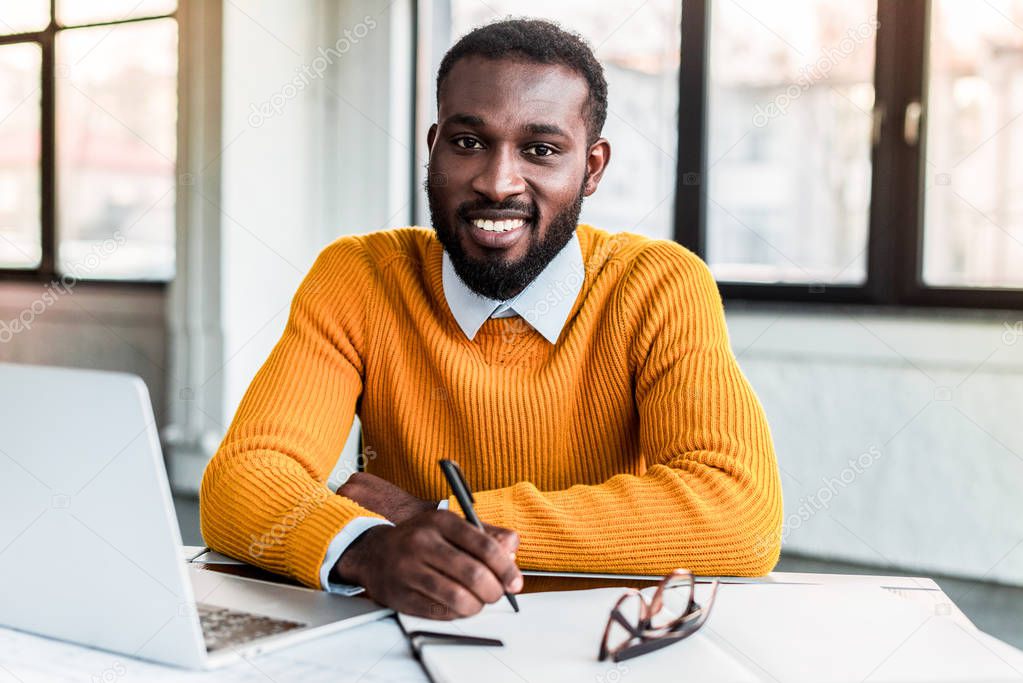 smiling african american businessman holding pen and looking at camera in office