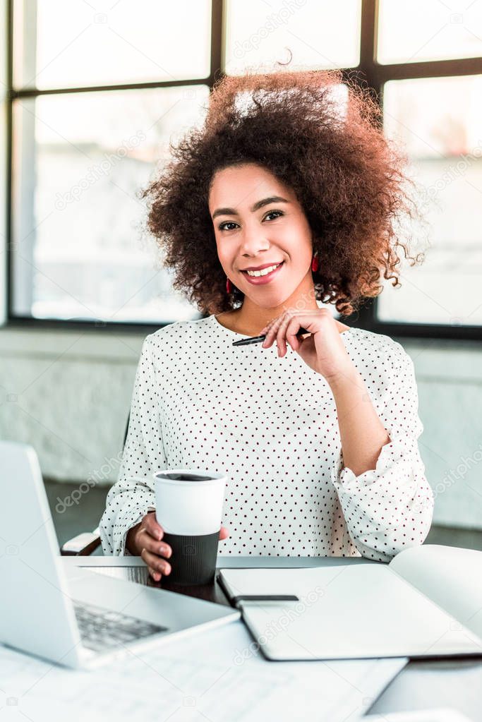 african american businesswoman holding cup of coffee and looking at camera in office