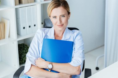attractive adult female doctor holding clipboard and looking at camera in office  clipart